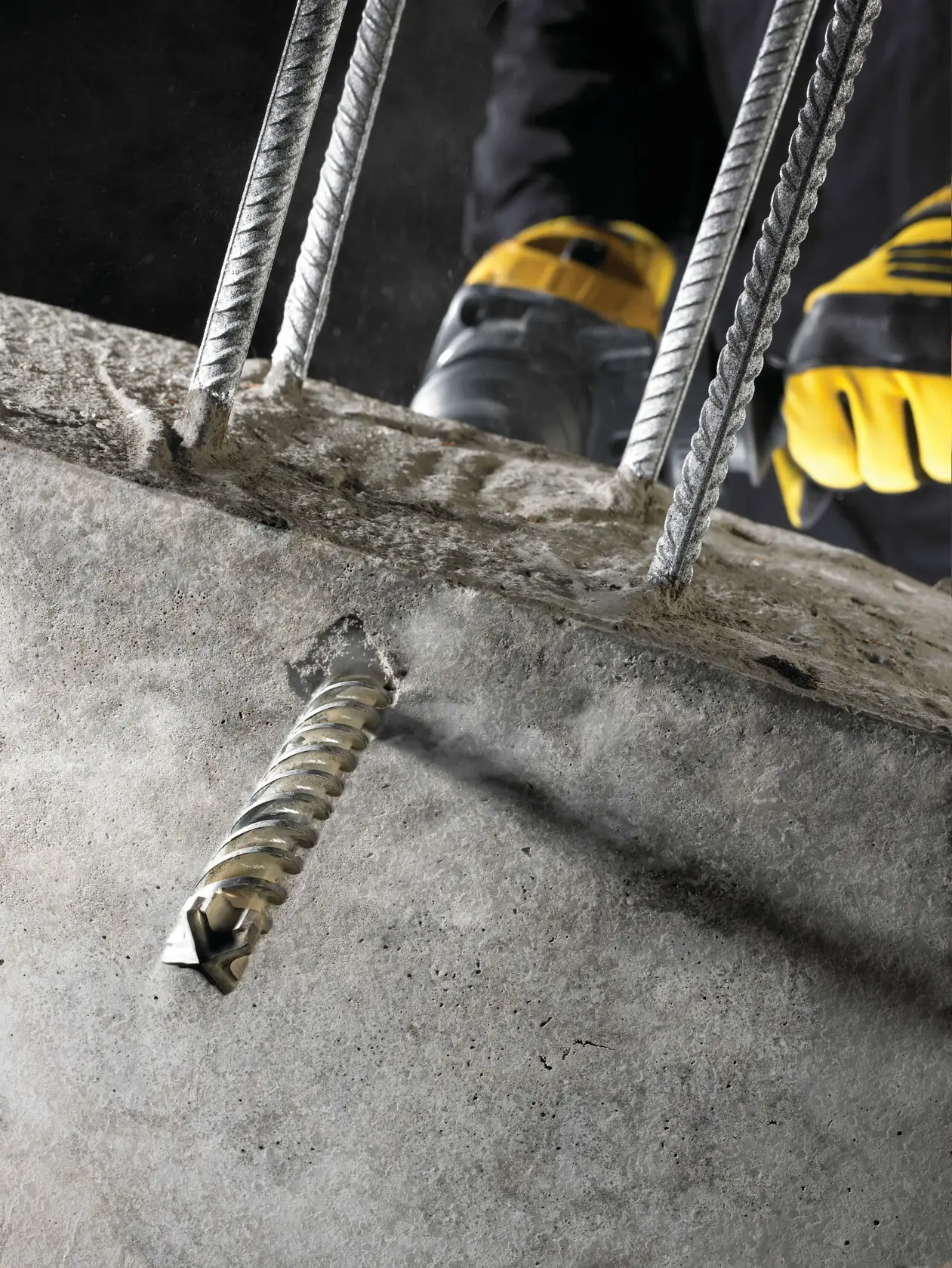 Close-up de uma broca DEWALT perfurando concreto, vergalhões e mãos com luvas visíveis.