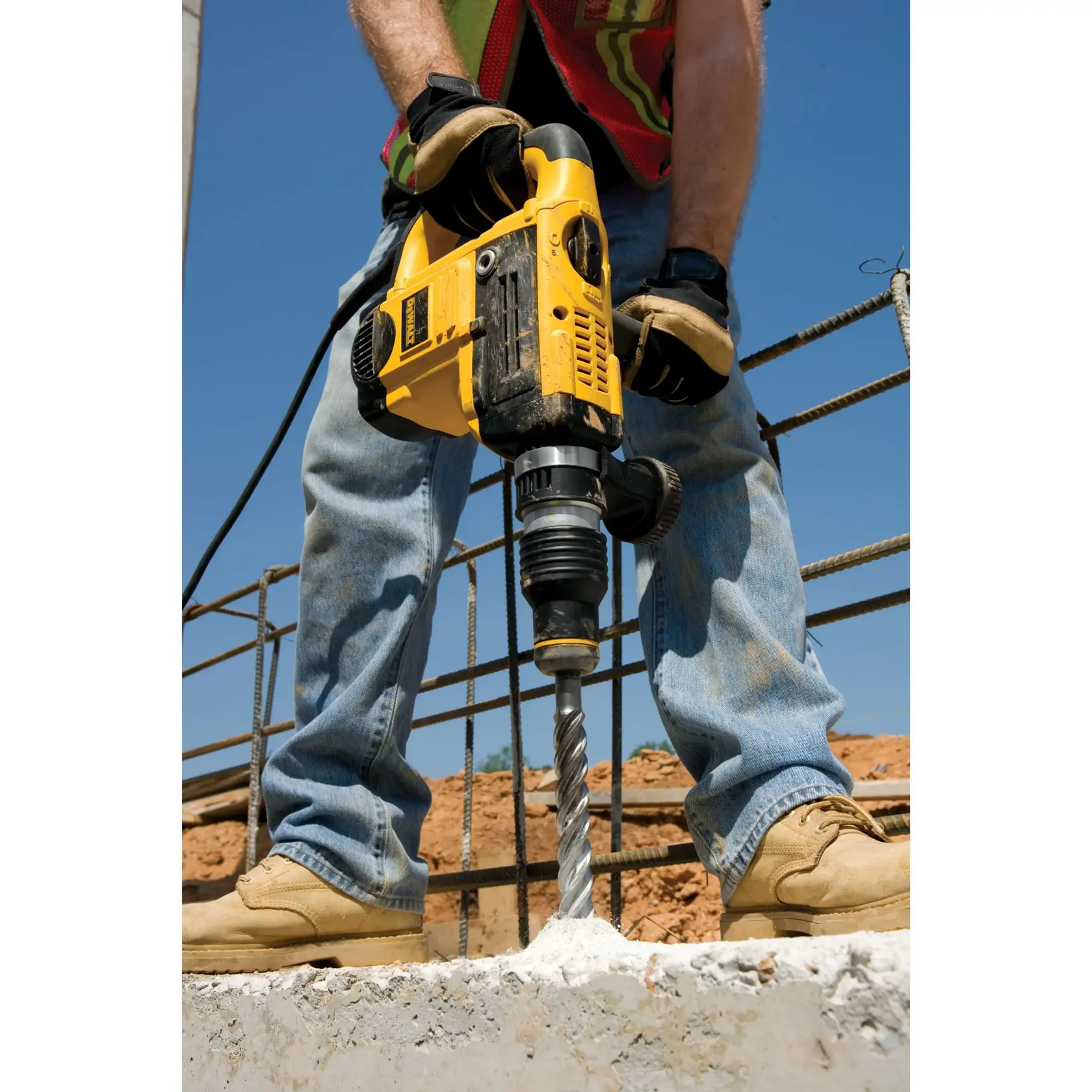 Pessoa perfura concreto com martelo perfurador DEWALT amarelo em obra, usando luvas e botas.