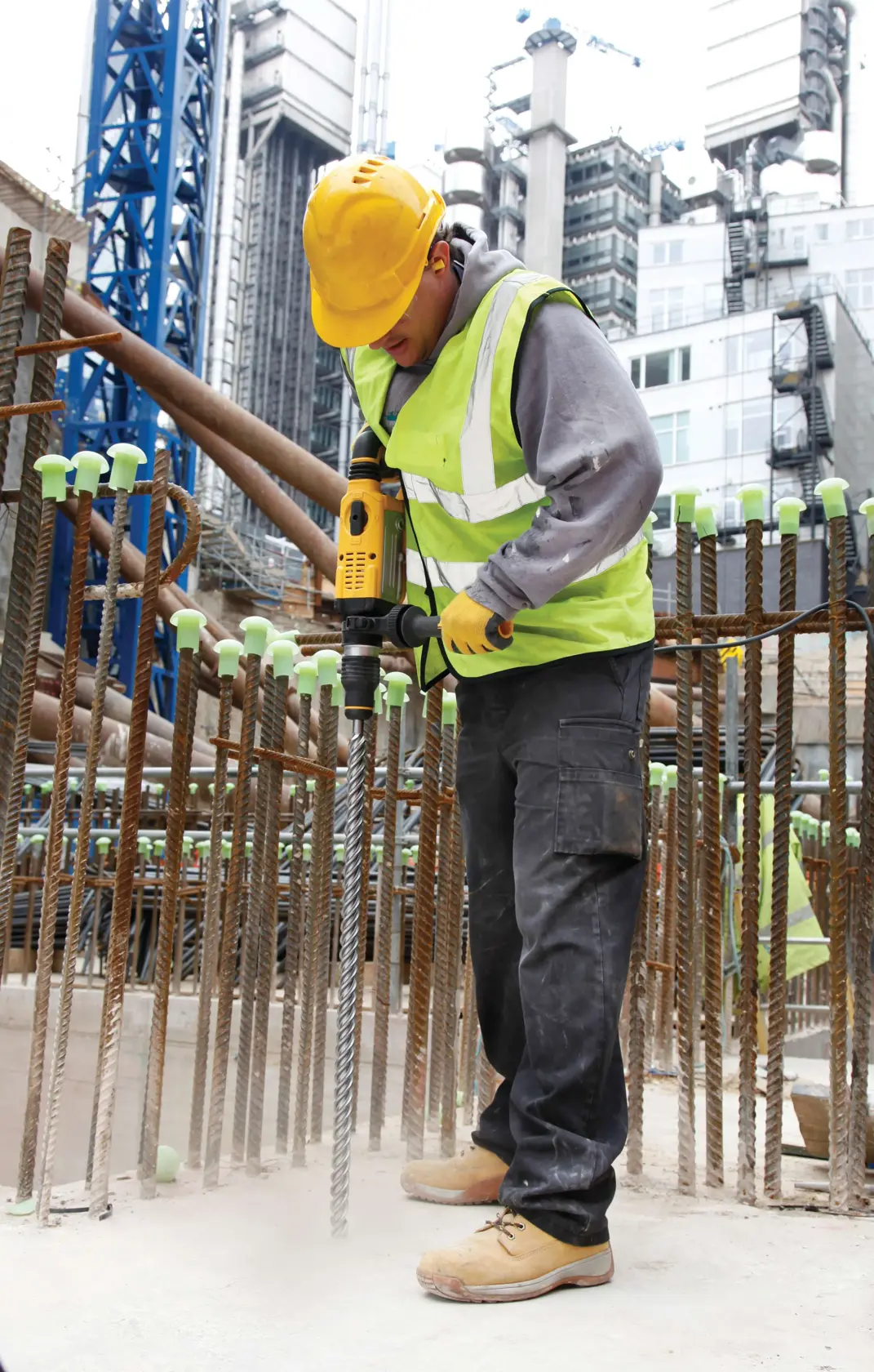 Trabalhador usa furadeira DEWALT na obra, cercado por vergalhões e usando equipamento de segurança.