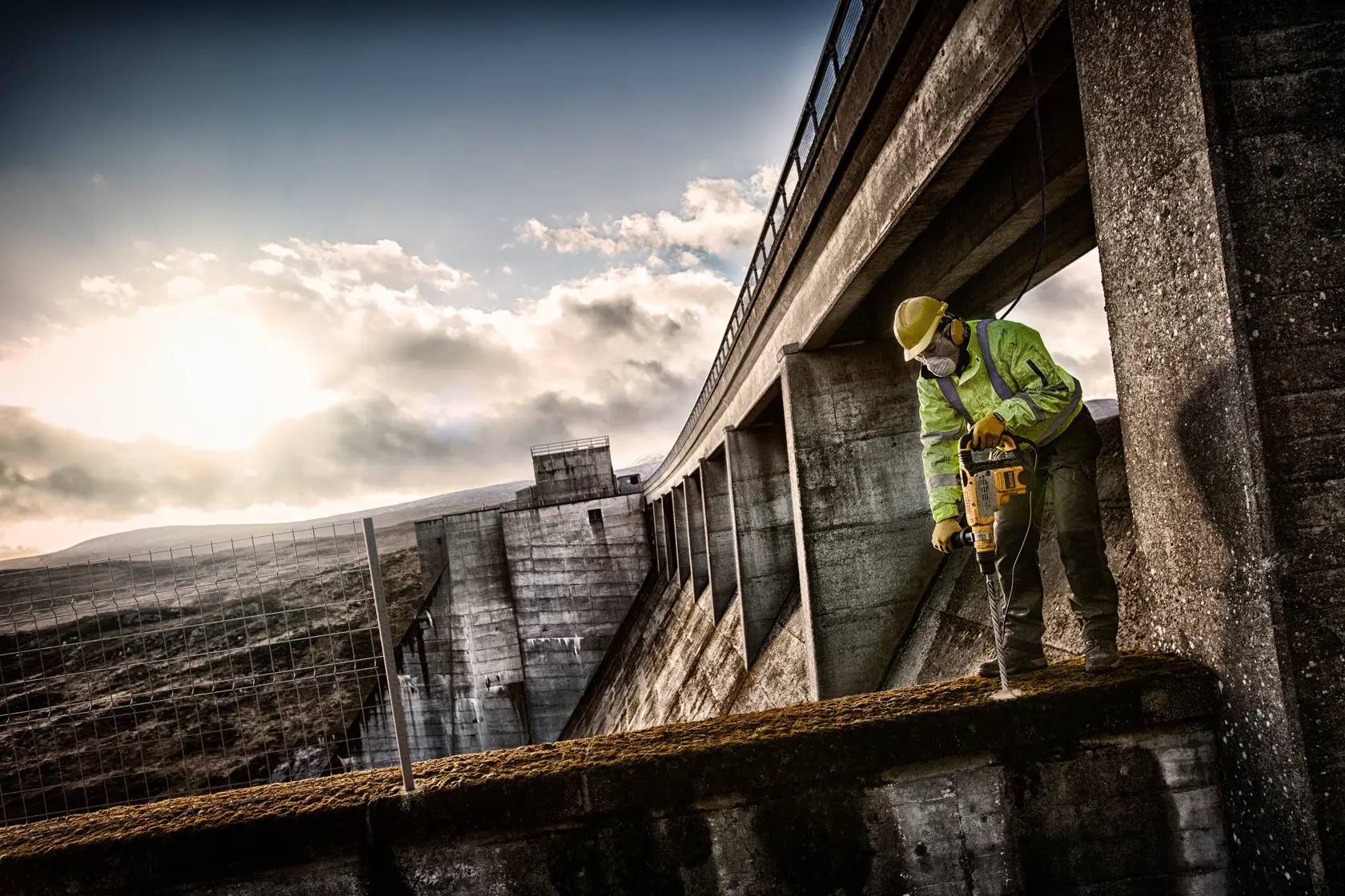 Trabajador con protección usa herramienta DEWALT para perforar concreto cerca de estructura exterior grande.