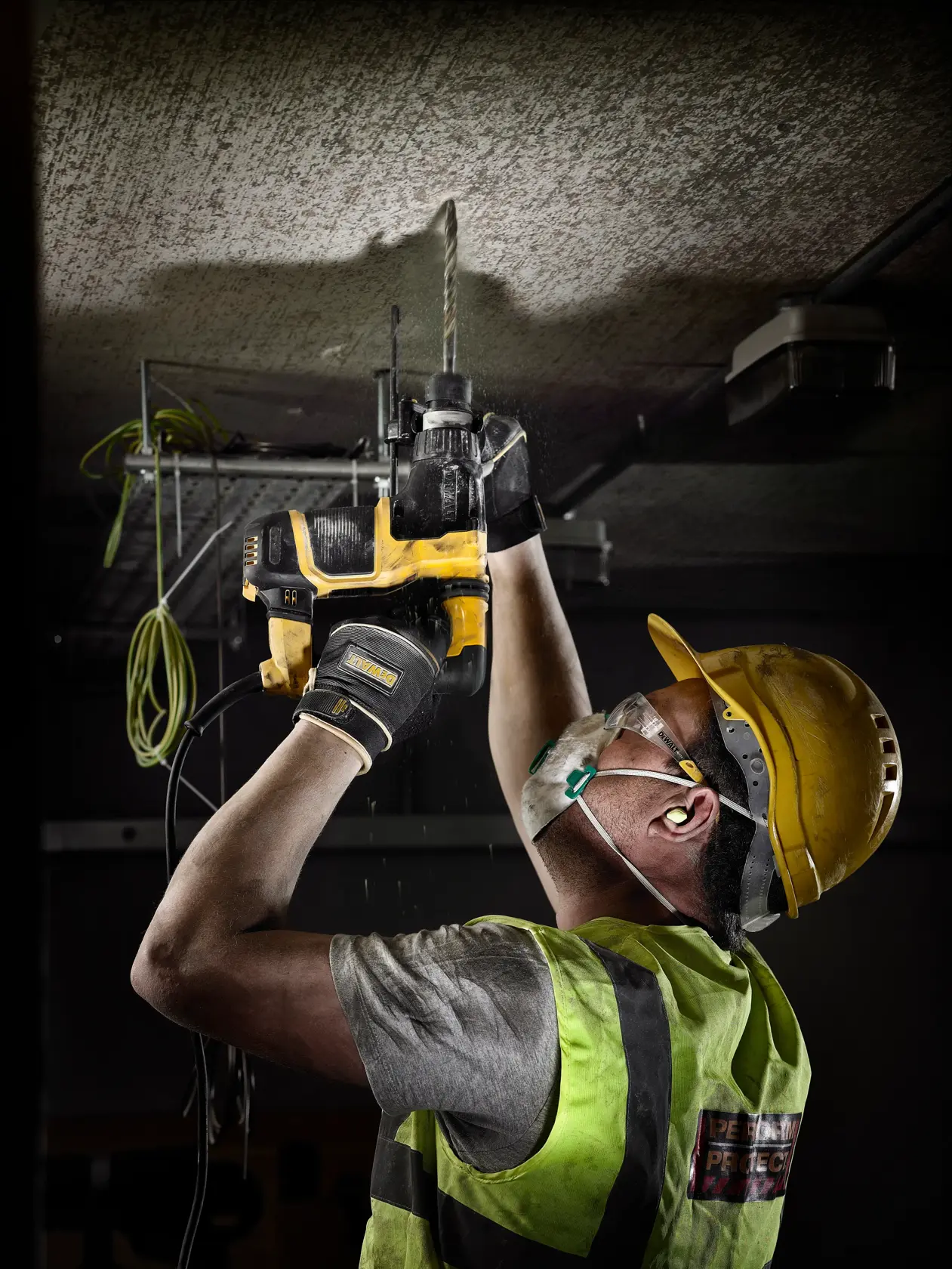 Um trabalhador da construção civil usando equipamentos de segurança utiliza uma furadeira elétrica DEWALT para perfurar o teto.