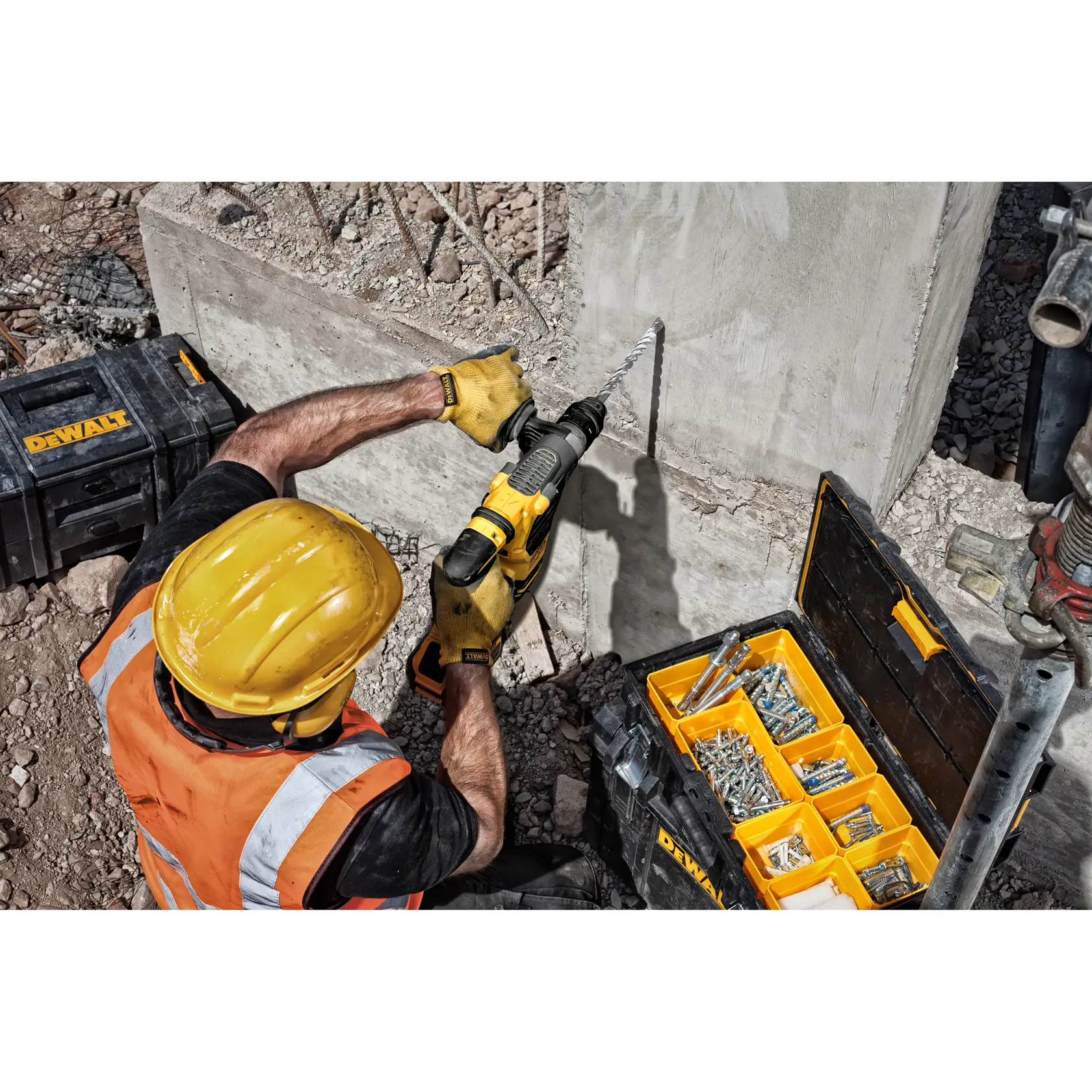 Trabajador perfora muro de concreto con herramienta DEWALT, casco y chaleco naranja, junto a cajas y fijaciones.