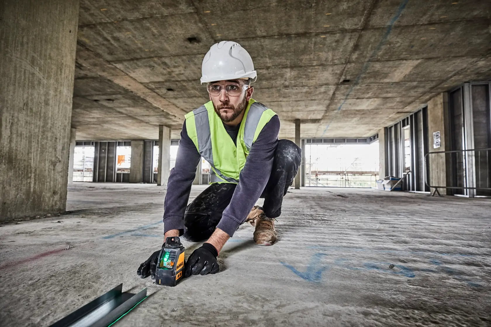 Um trabalhador usando capacete, colete de segurança e luvas está ajoelhado no piso de um canteiro de obras e opera uma ferramenta de laser de linha cruzada verde DEWALT DW088CG para nivelamento e alinhamento.