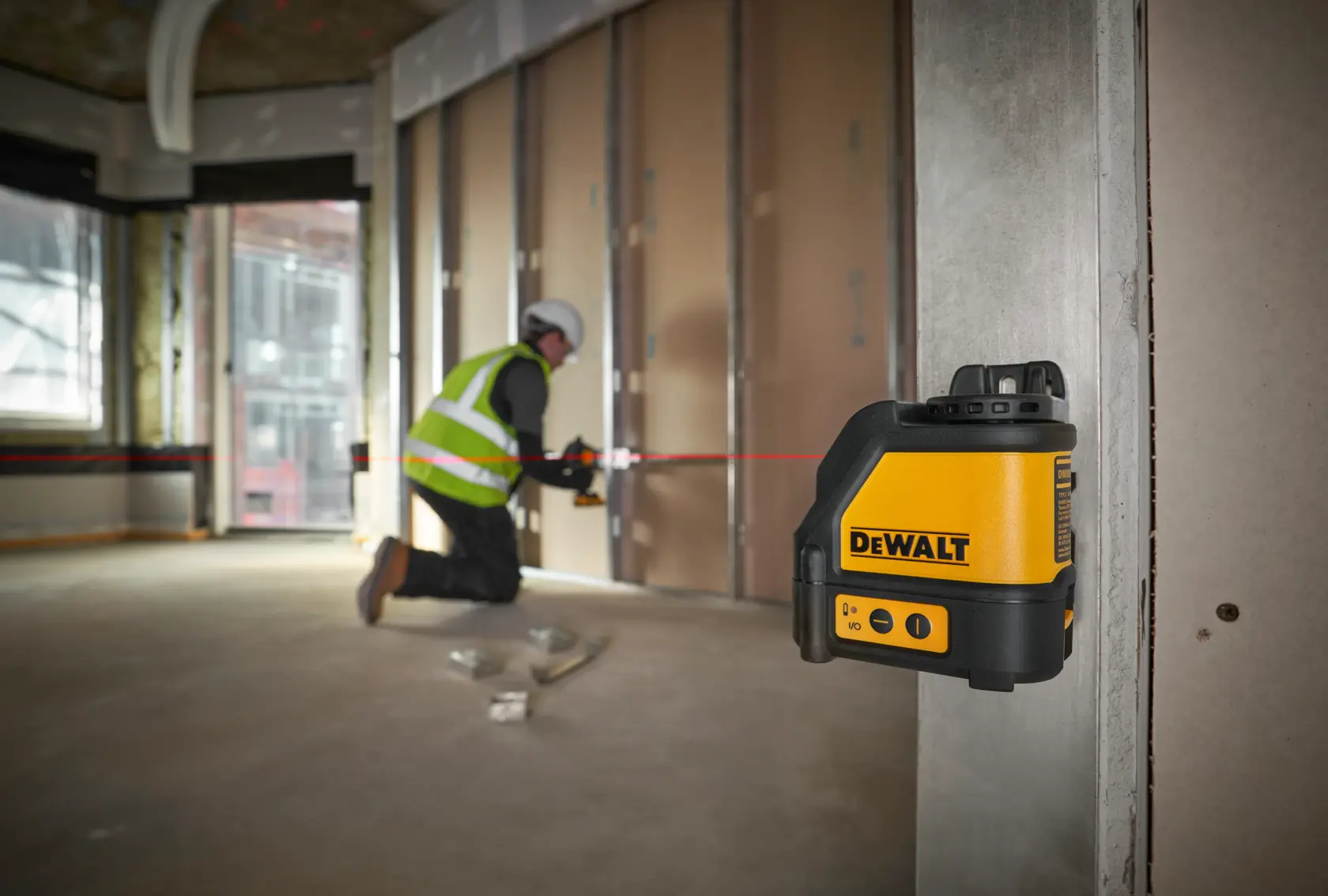 Um nível a laser DEWALT DW088K está fixado em uma coluna de metal, projetando uma linha horizontal vermelha pelo interior de um local de construção. Ao fundo, um trabalhador usando colete de segurança e capacete está alinhando materiais ao longo da linha do laser.