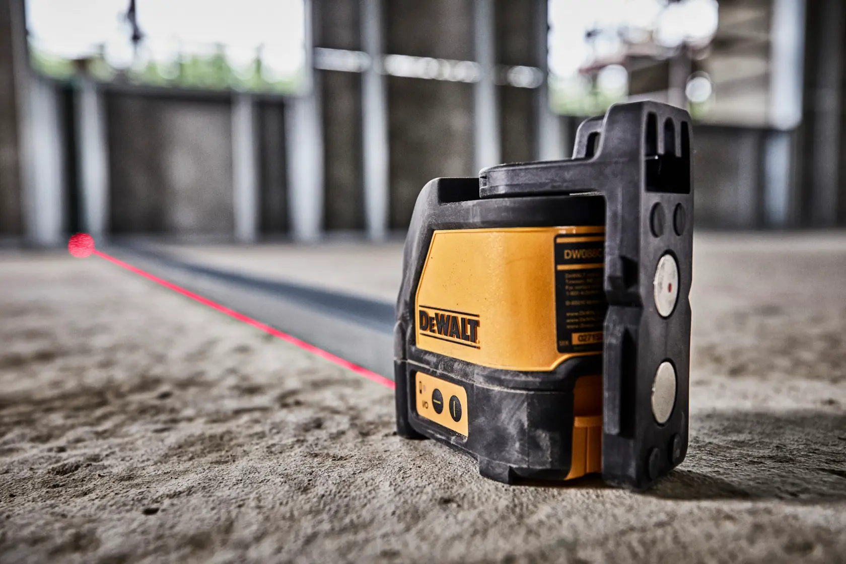 Uma pessoa usando capacete, colete de segurança e luvas está ajoelhada no chão de um canteiro de obras, usando um nivelador a laser DEWALT DW088K. O dispositivo projeta uma linha de laser vermelho visível para alinhamento. O ambiente mostra um edifício inacabado com colunas de concreto e janelas abertas.