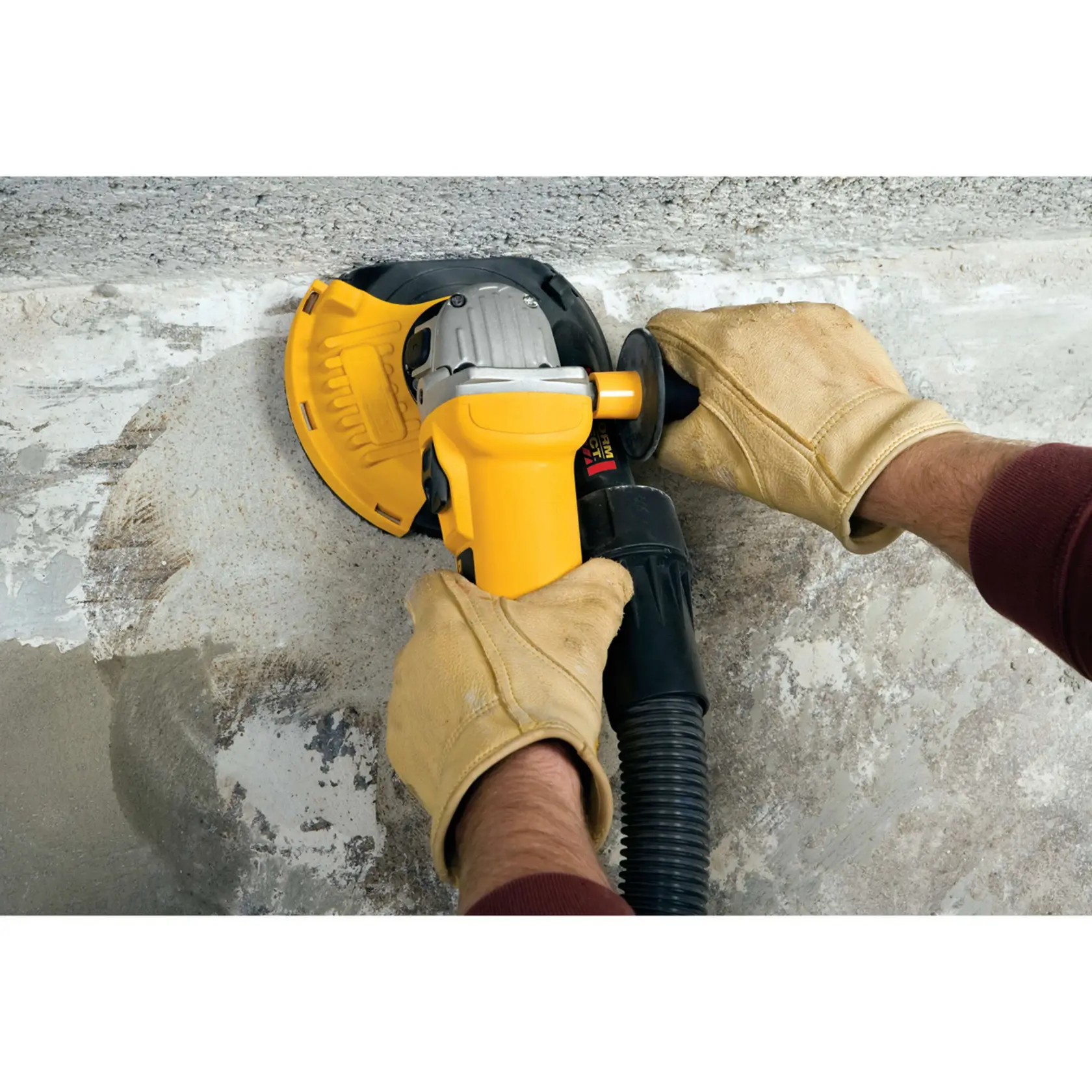 Um trabalhador da construção civil usando capacete amarelo, luvas e máscara facial está utilizando uma esmerilhadeira angular DEWALT com proteção contra poeira em um piso de concreto. Um aspirador DEWALT está conectado à ferramenta para coletar poeira. A cena acontece dentro de um prédio em construção, com outros trabalhadores ao fundo.