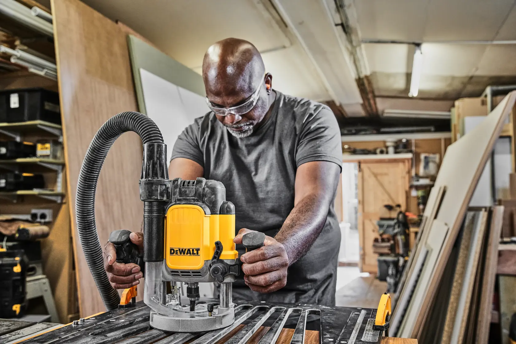 Uma pessoa está usando uma tupia DEWALT DWE625 em uma oficina de marcenaria. A ferramenta está sobre uma bancada com peças de madeira e uma mangueira de aspiração conectada. A oficina contém vários materiais e equipamentos.