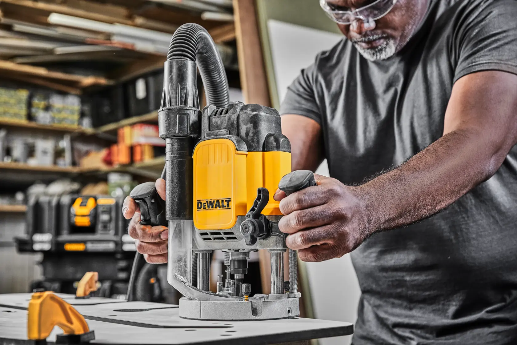 Uma pessoa utiliza uma tupia DEWALT DWE625 em uma bancada de trabalho em uma oficina. A ferramenta amarela e preta está equipada com uma mangueira de extração de pó. Outros equipamentos DEWALT e prateleiras com suprimentos são visíveis ao fundo.