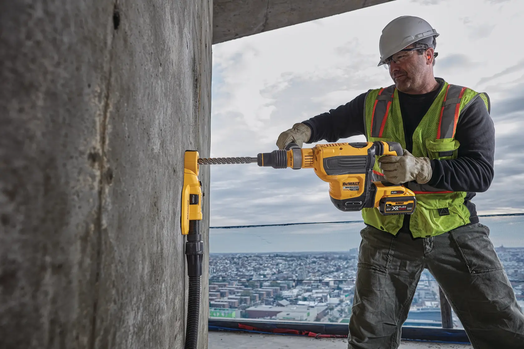 Um trabalhador da construção civil usa um martelete rotativo DEWALT conectado a uma ferramenta de extração de pó para perfurar uma parede de concreto em um canteiro de obras de edifício alto.