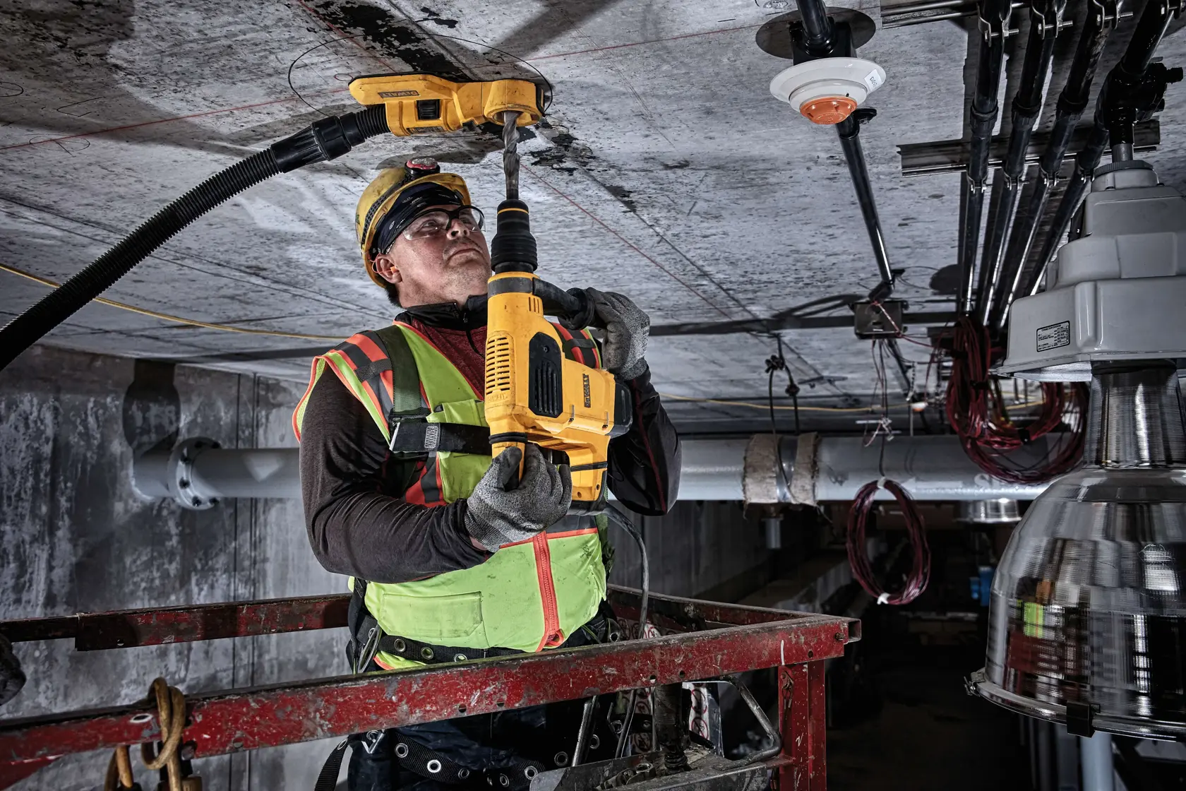 Um trabalhador usando colete de segurança verde e capacete está utilizando um sistema de extração de pó DEWALT DWH050 acoplado a uma furadeira amarela, perfurando o teto de concreto em um ambiente industrial.
