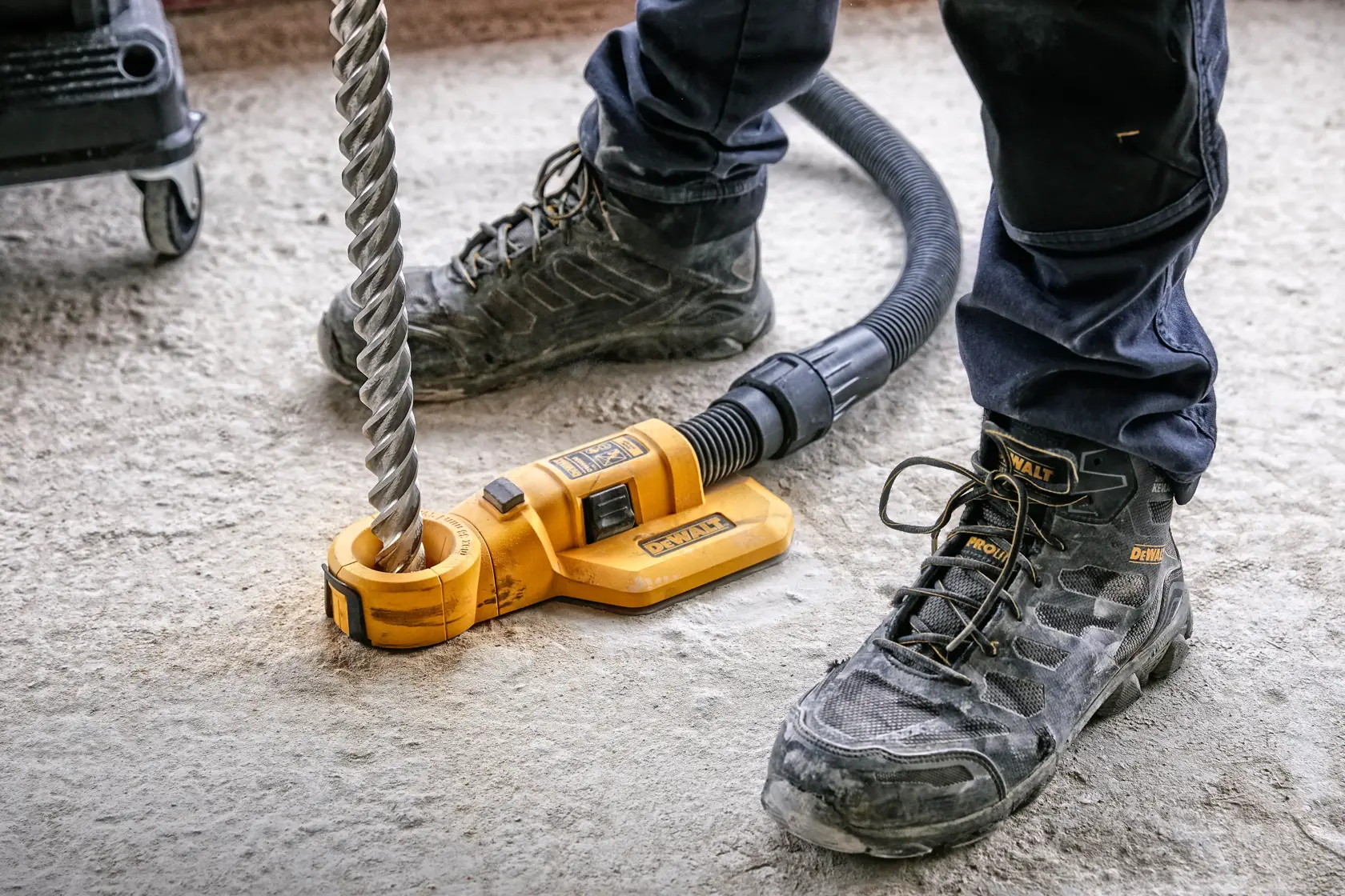 Close-up de um acessório de extração de poeira DEWALT DWH050 conectado a uma broca grande e mangueira, sendo utilizado em uma superfície de concreto por alguém usando botas de segurança e calça de trabalho.