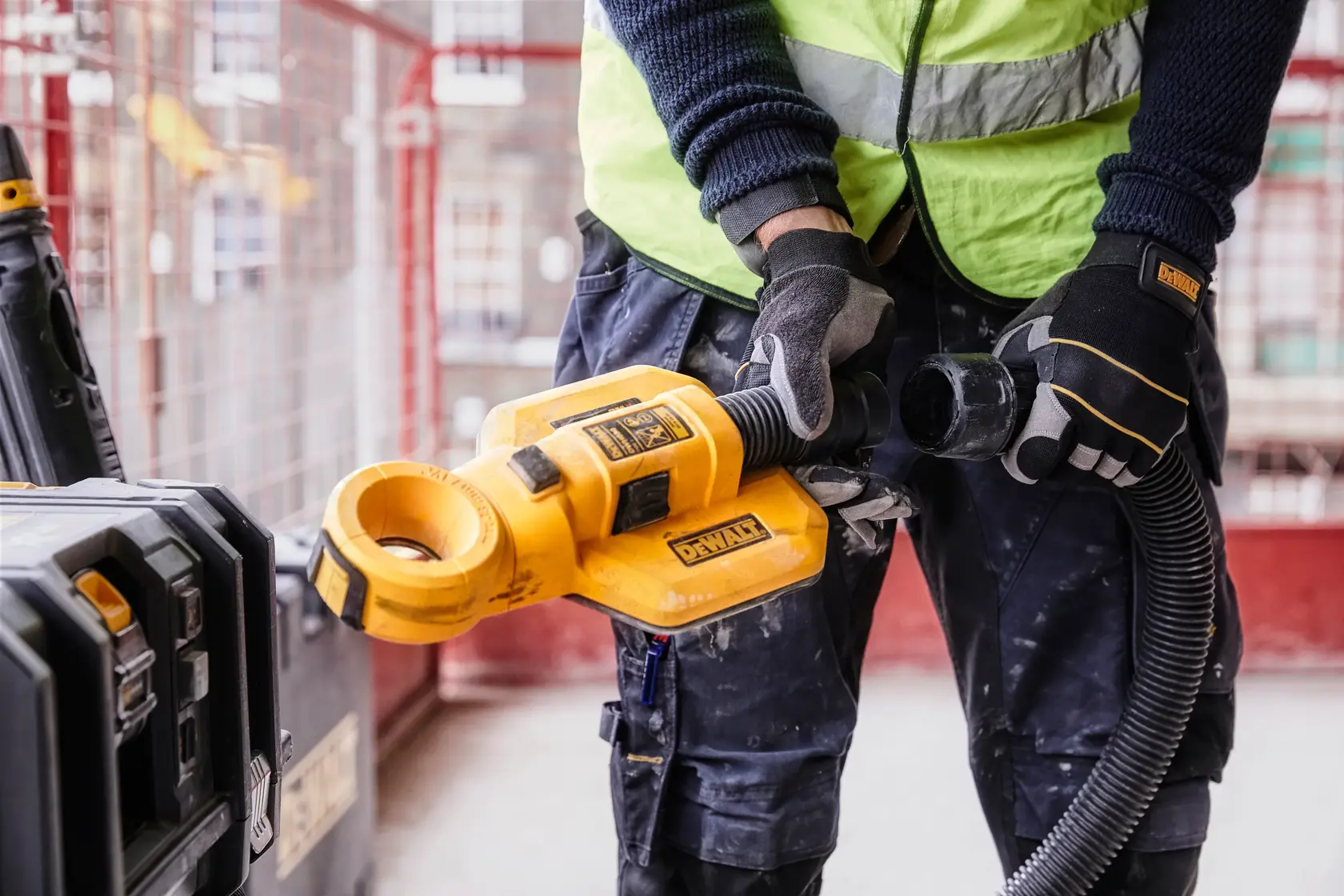 Uma pessoa com luvas de proteção e colete refletivo utiliza um acessório de coleta de poeira DEWALT DWH050 em um canteiro de obras.