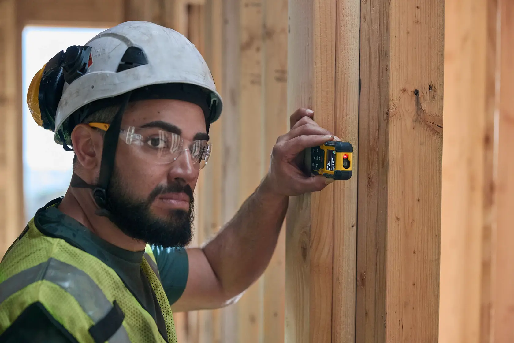 Um trabalhador da construção usando capacete de segurança e colete refletivo está utilizando um medidor de distância a laser DEWALT DWHT77200 em uma viga de madeira dentro de um edifício em construção. O rosto do trabalhador está encoberto.