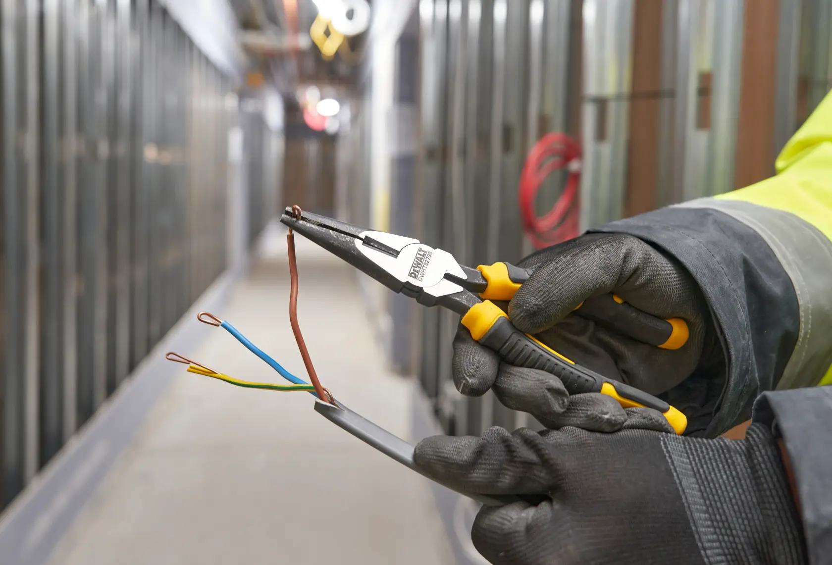Manos con guantes usan alicates DEWALT para doblar cables eléctricos en pasillo en construcción.