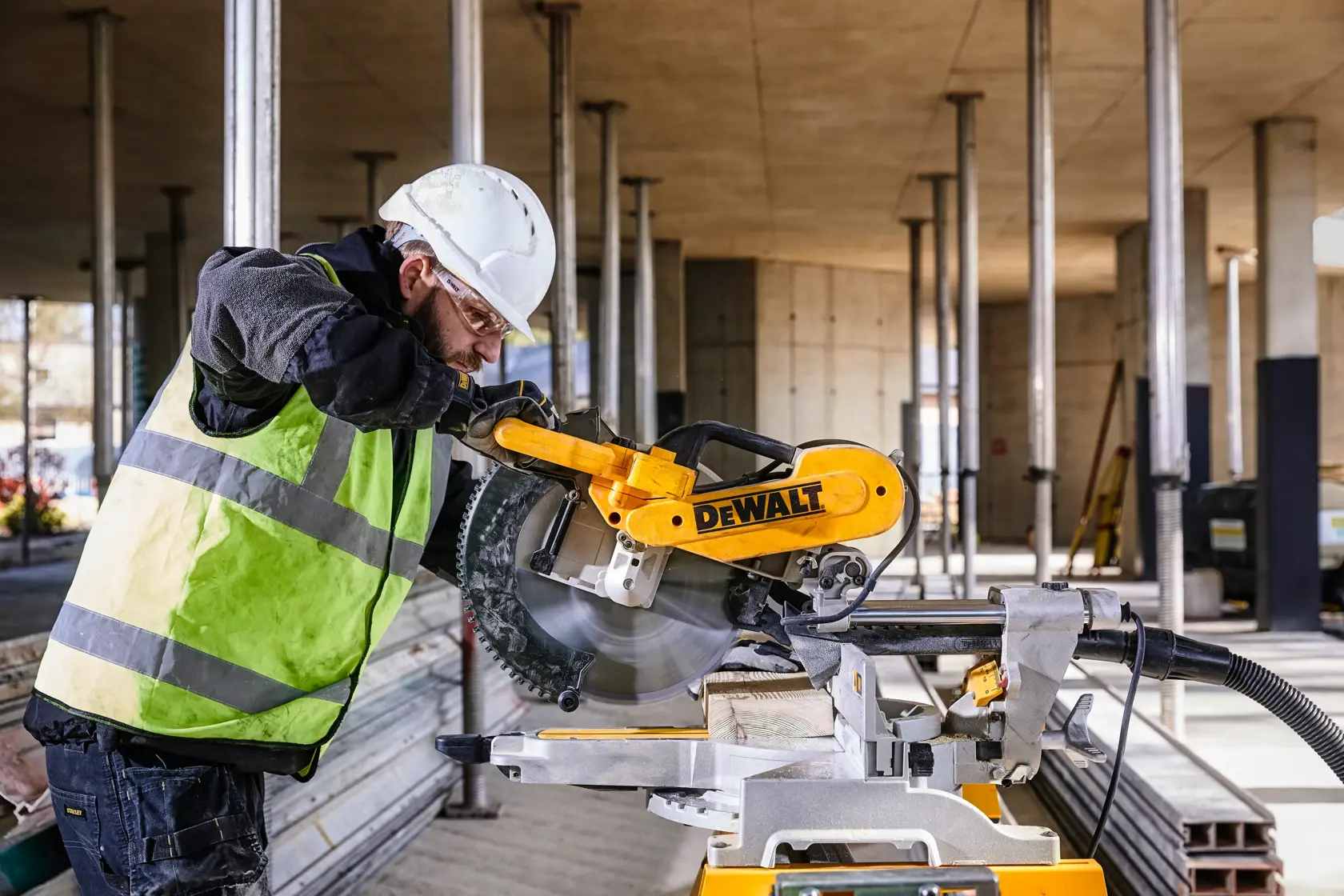 Uma pessoa com capacete e colete de segurança está operando uma serra de meia-esquadria DEWALT DWS780 em um canteiro de obras, com suportes metálicos e materiais de construção visíveis ao fundo.