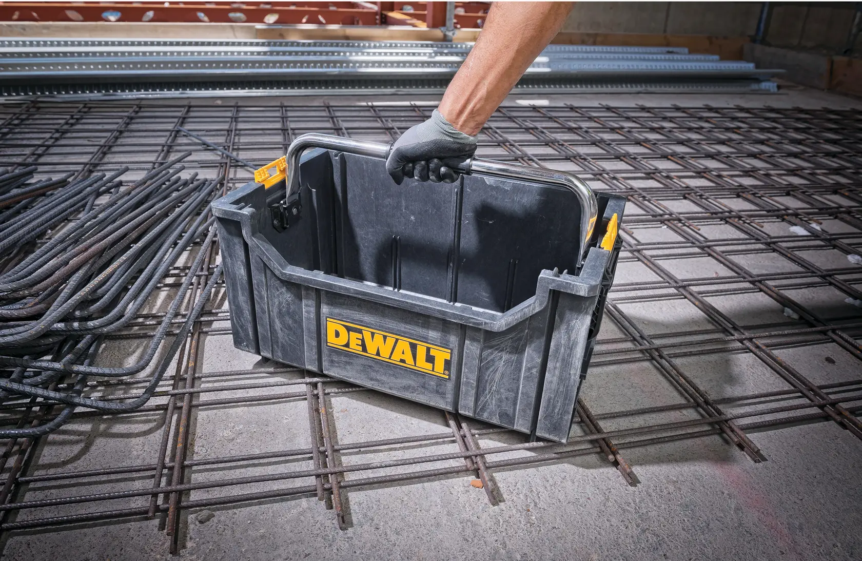 Pessoa ergue uma caixa de ferramentas DEWALT de plástico com alça sobre grade de metal em obra.
