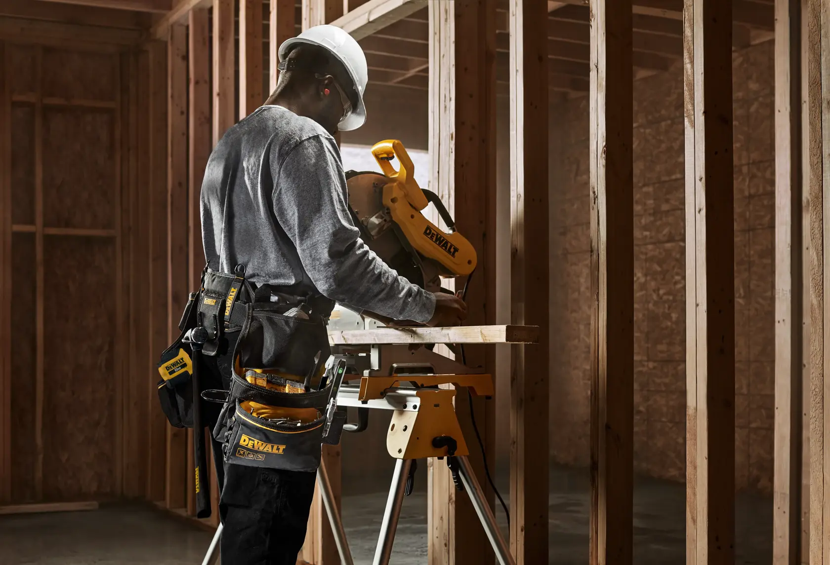 Persona con casco y cinturón de herramientas operando una sierra de inglete DEWALT en un sitio de construcción con estructura de madera.