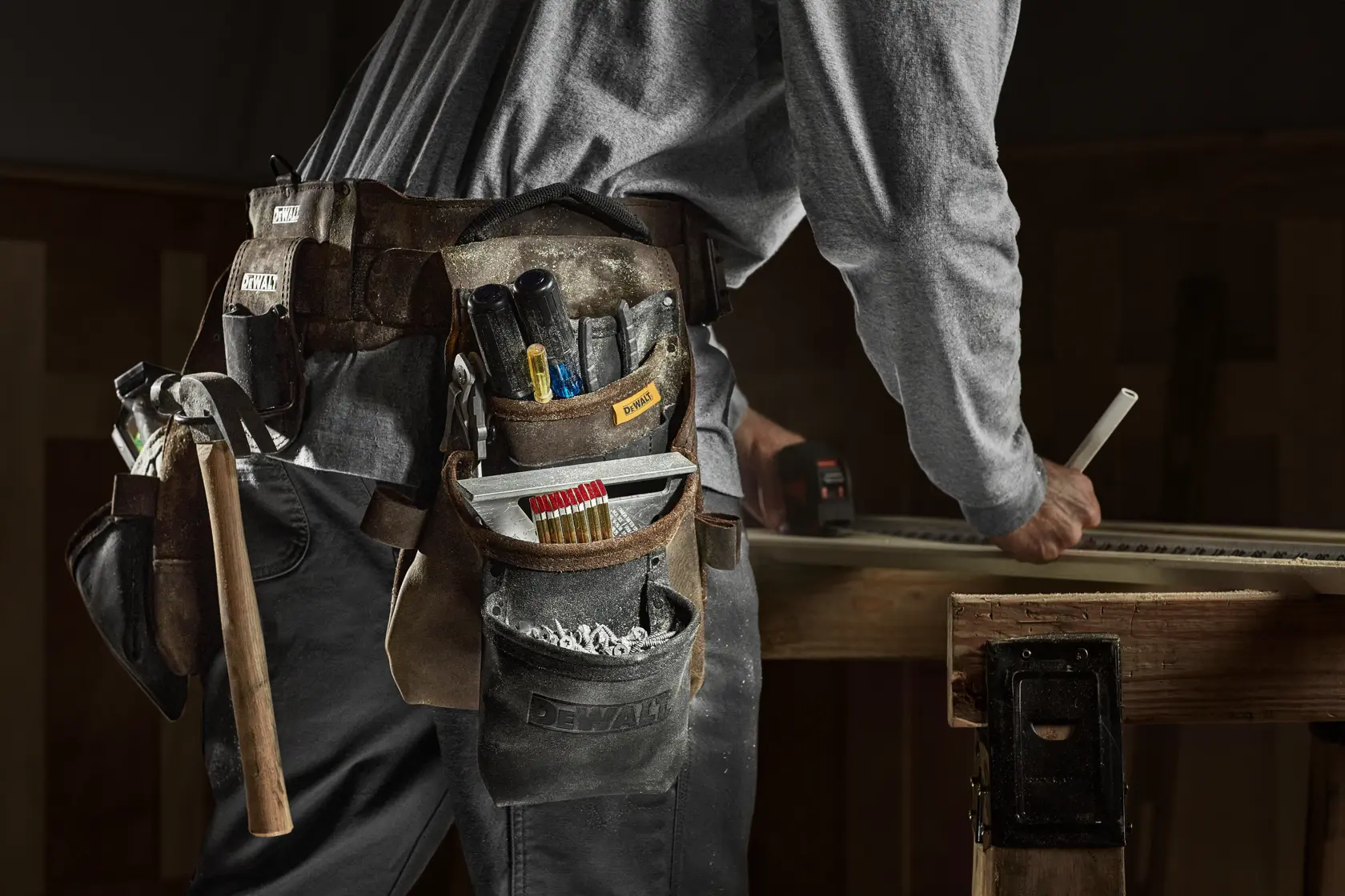 Primer plano de una persona que lleva un cinturón de herramientas DEWALT cargado con varios instrumentos como un martillo, lápices y destornilladores. La persona está trabajando en una mesa de madera marcando una pieza de madera.