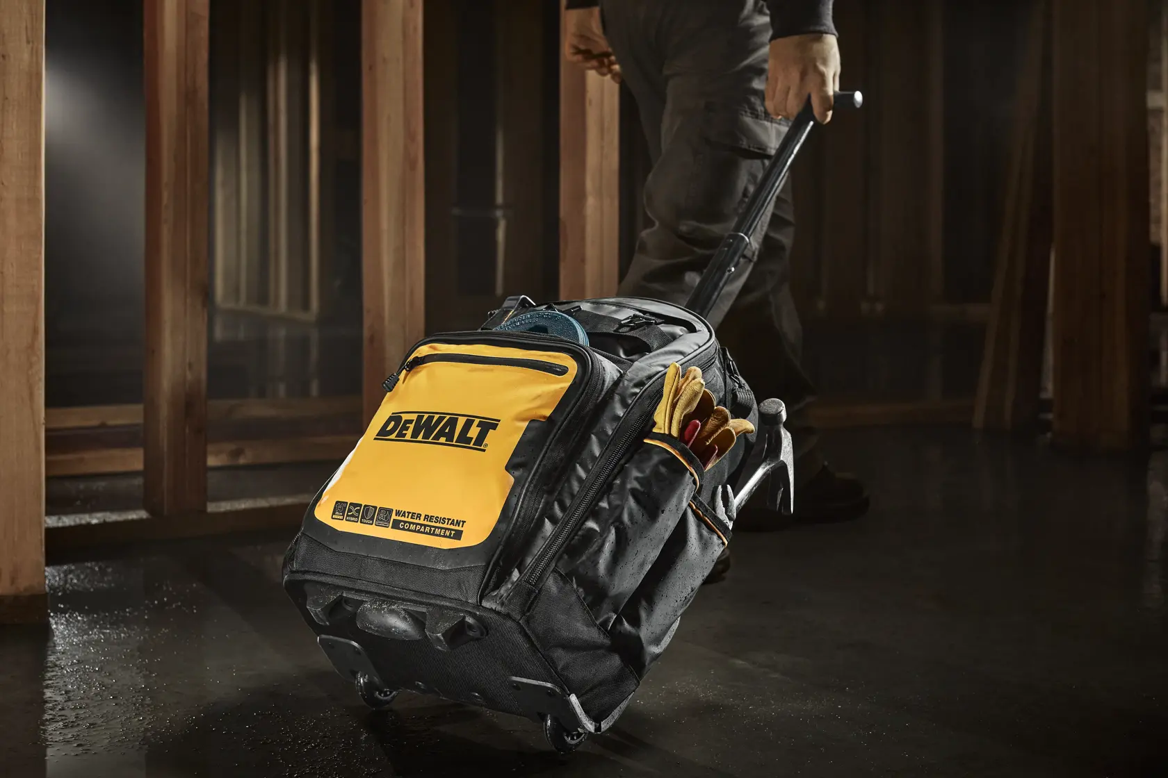 Uma bolsa de ferramentas DEWALT preta e amarela com rodas e alça extensível, sendo puxada por uma pessoa em um ambiente de trabalho. A bolsa possui bolsos externos contendo luvas de trabalho e outros acessórios para ferramentas.