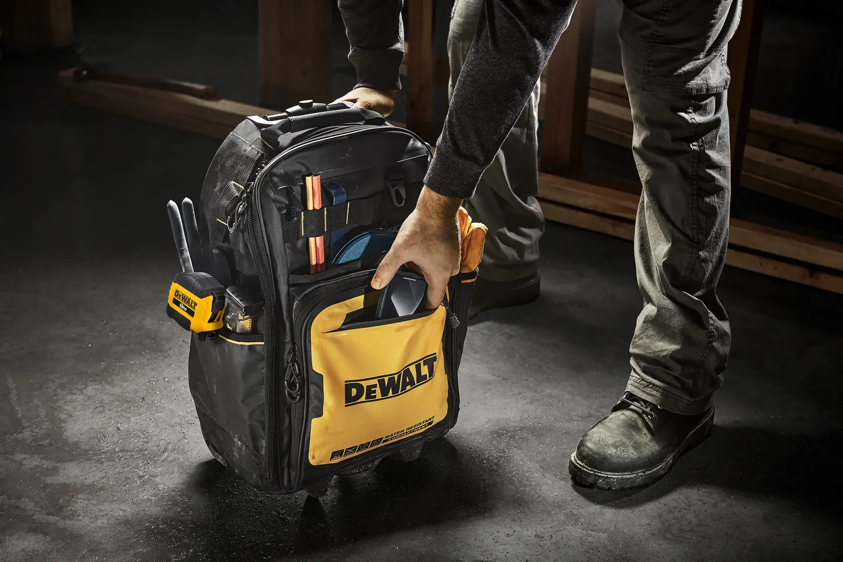 Uma pessoa segura uma mochila de ferramentas da marca DEWALT, cheia de várias ferramentas e equipamentos. A mochila é amarela e preta e está posicionada no chão de uma oficina.