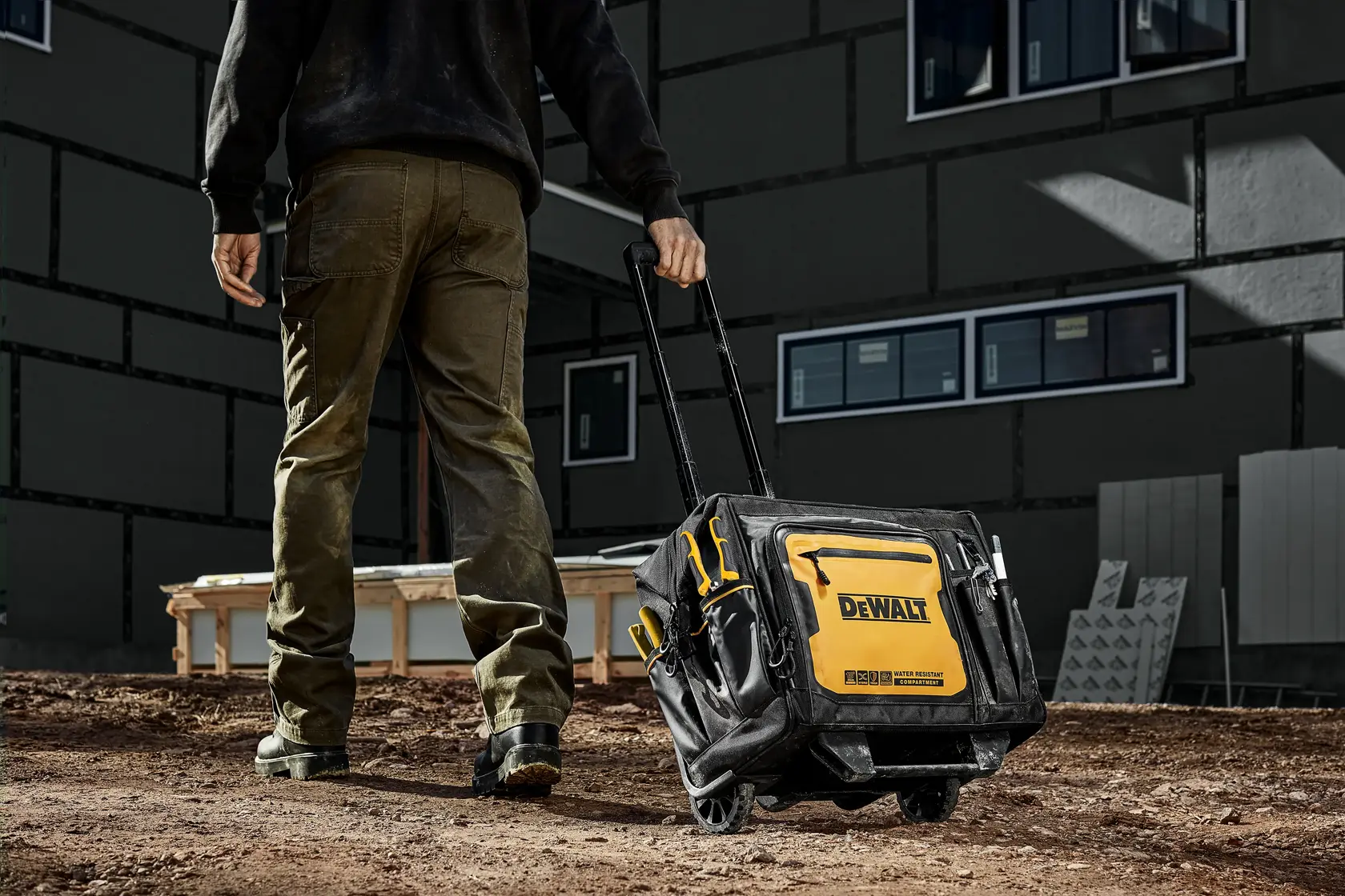 Uma pessoa está puxando uma mala de ferramentas DEWALT com rodas e alça telescópica em um canteiro de obras de terra. A mala é preta e amarela, com o logotipo DEWALT e vários compartimentos para armazenamento.