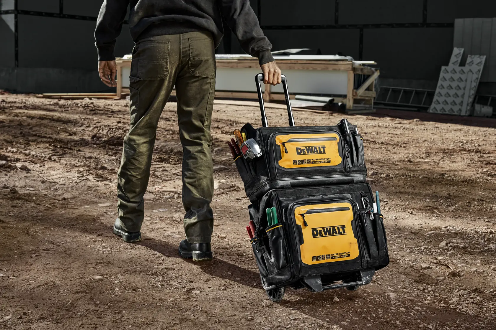 Uma pessoa está puxando uma bolsa de ferramentas com rodas DEWALT em um local de construção. A bolsa é preta e amarela, com vários bolsos e compartimentos, e está cheia de diversas ferramentas.