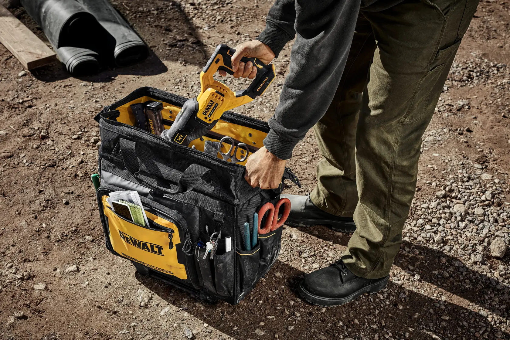 Uma pessoa coloca uma ferramenta elétrica amarela DEWALT em uma bolsa de ferramentas preta e amarela DEWALT cheia de vários instrumentos e acessórios, sobre chão de terra em um local de trabalho ao ar livre.
