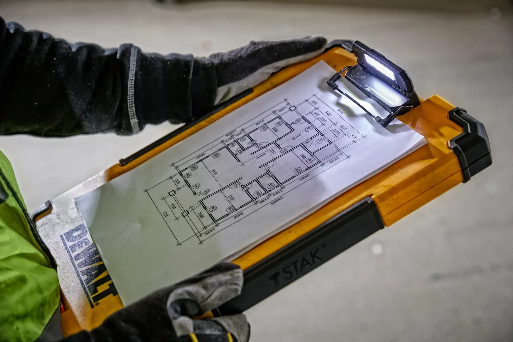 Una persona con guantes sostiene un portapapeles DEWALT TSTAK con luz LED integrada, mostrando un plano técnico sujetado en la pinza.