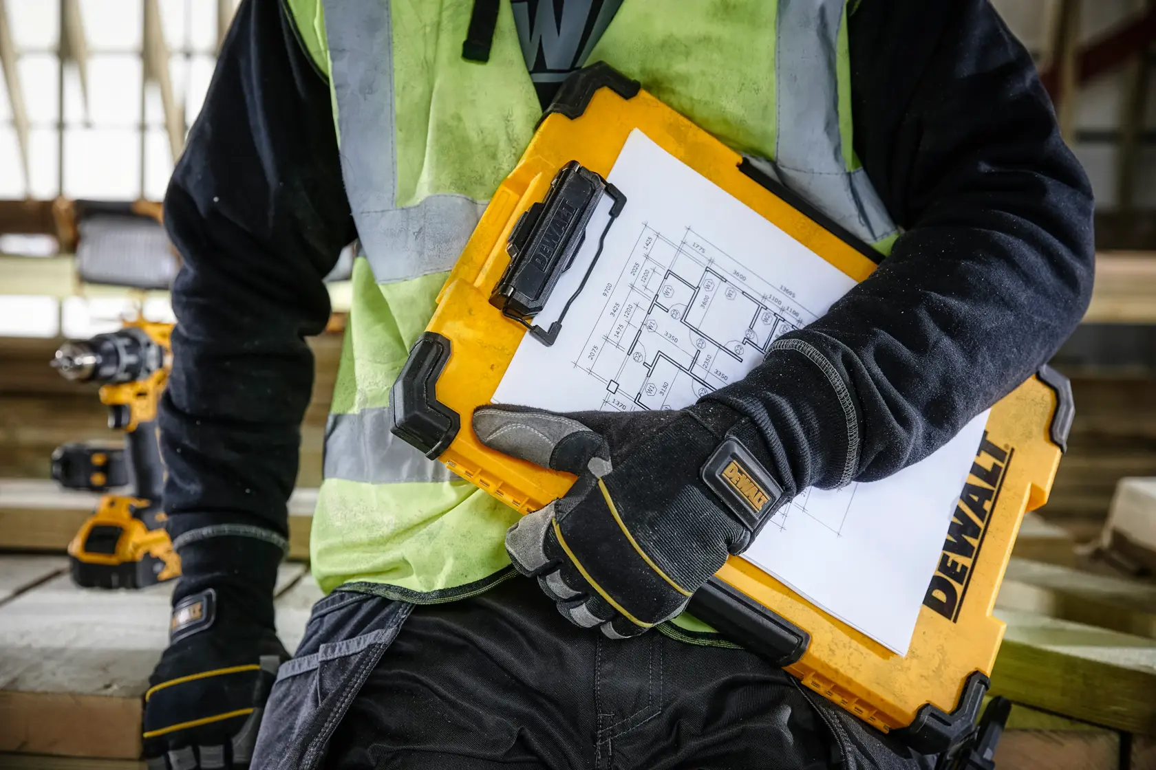 Una persona con guantes de trabajo y chaleco reflectante sostiene un portapapeles amarillo DEWALT con planos arquitectónicos adjuntos, en una obra de construcción.