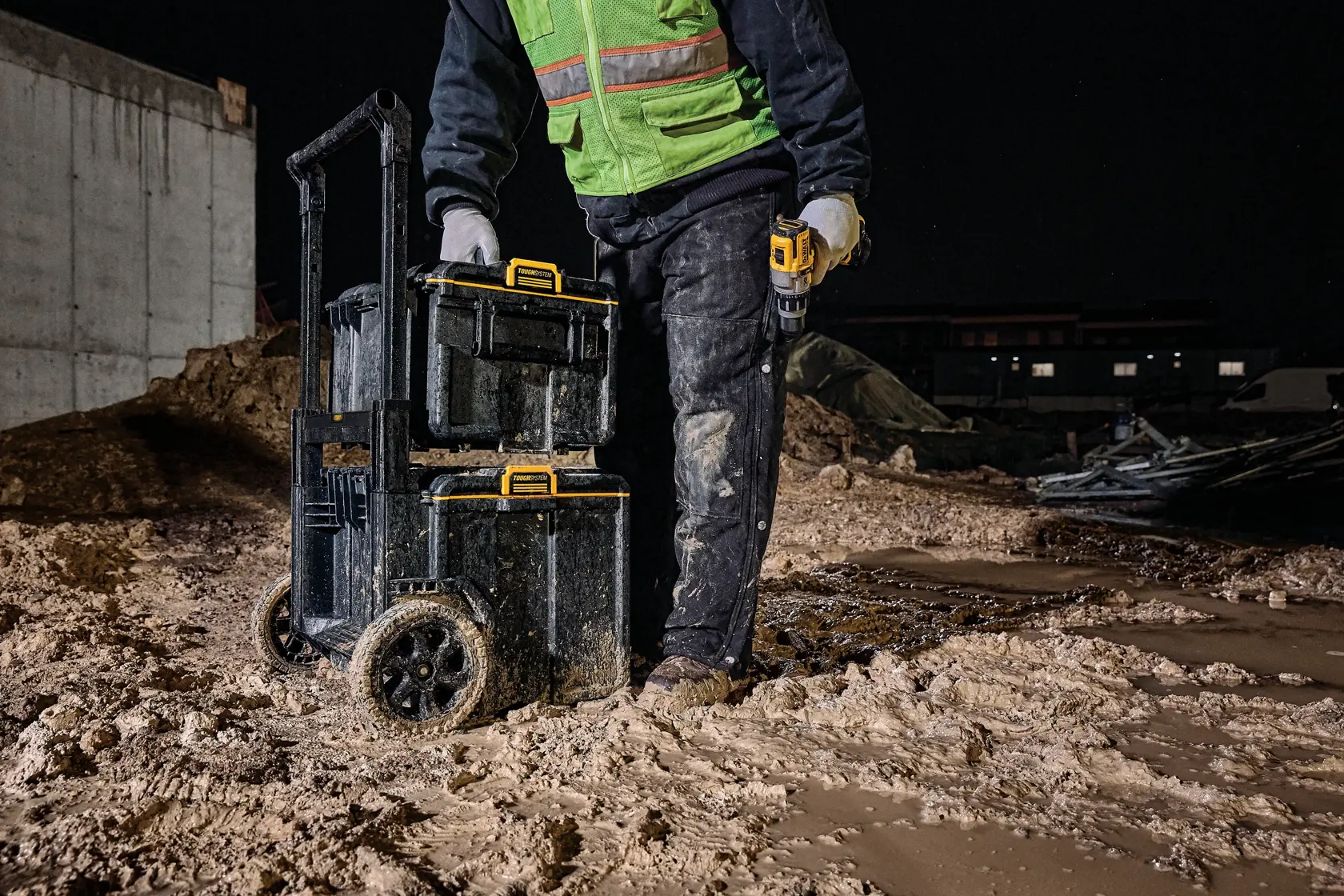 Persona con caja de herramientas DEWALT con ruedas en obra fangosa nocturna, con equipo de seguridad y taladro.