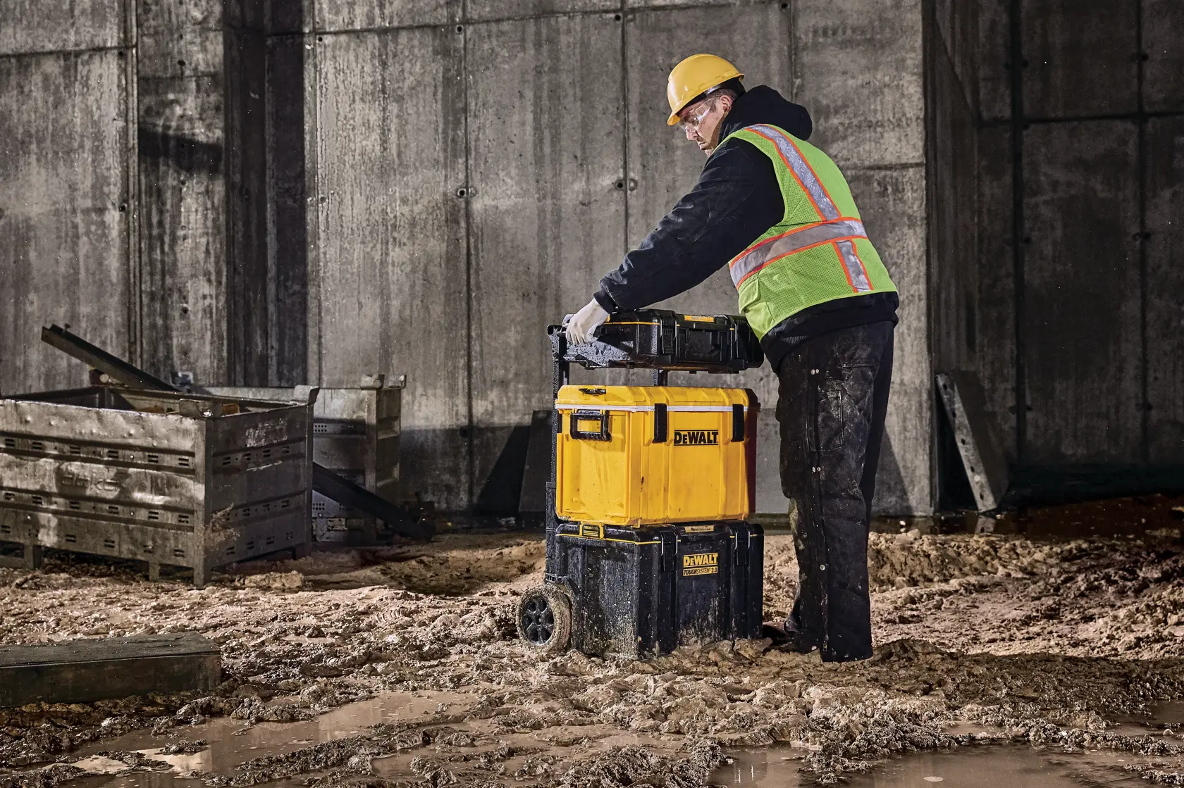 Trabajador mueve caja de herramientas DEWALT con ruedas en obra fangosa junto a cajas de madera.