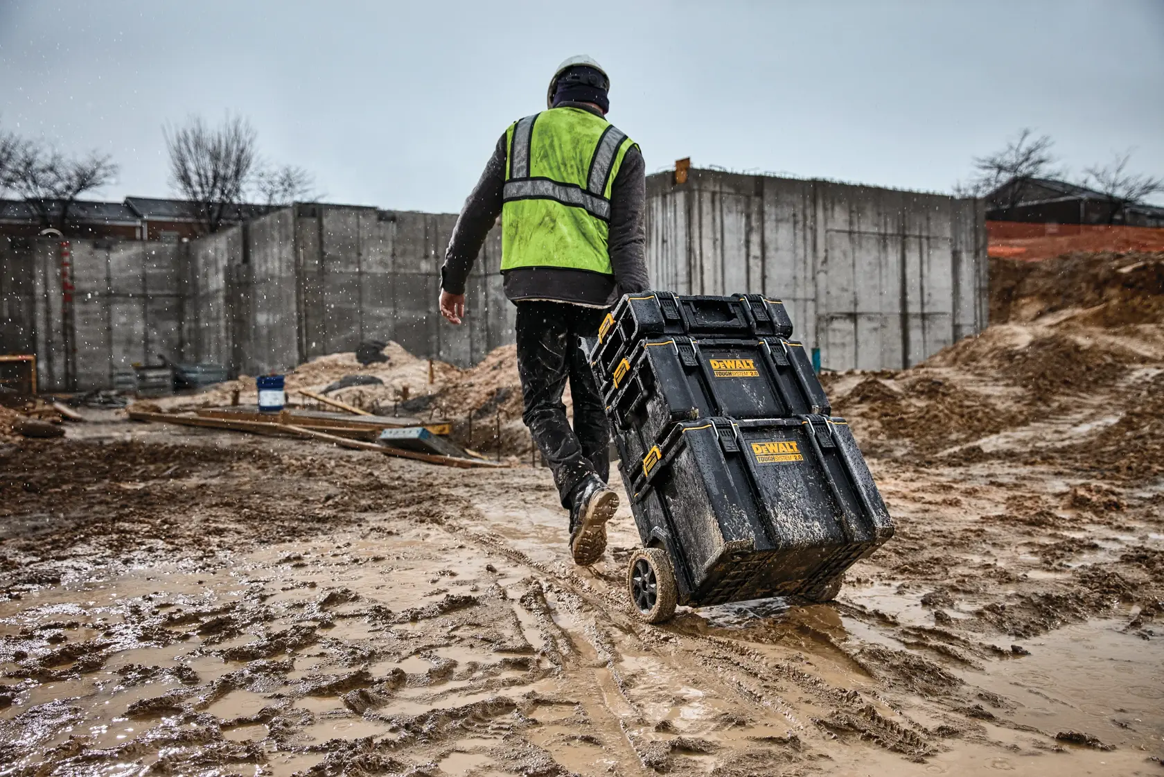 Trabajador arrastra cajas de herramientas DEWALT con ruedas en una obra fangosa, con chaleco de seguridad.