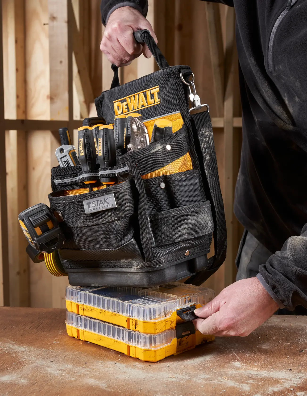 Bolsa de ferramentas DEWALT com ferramentas sobre organizadores amarelos, segurada por pessoa com roupa de trabalho.