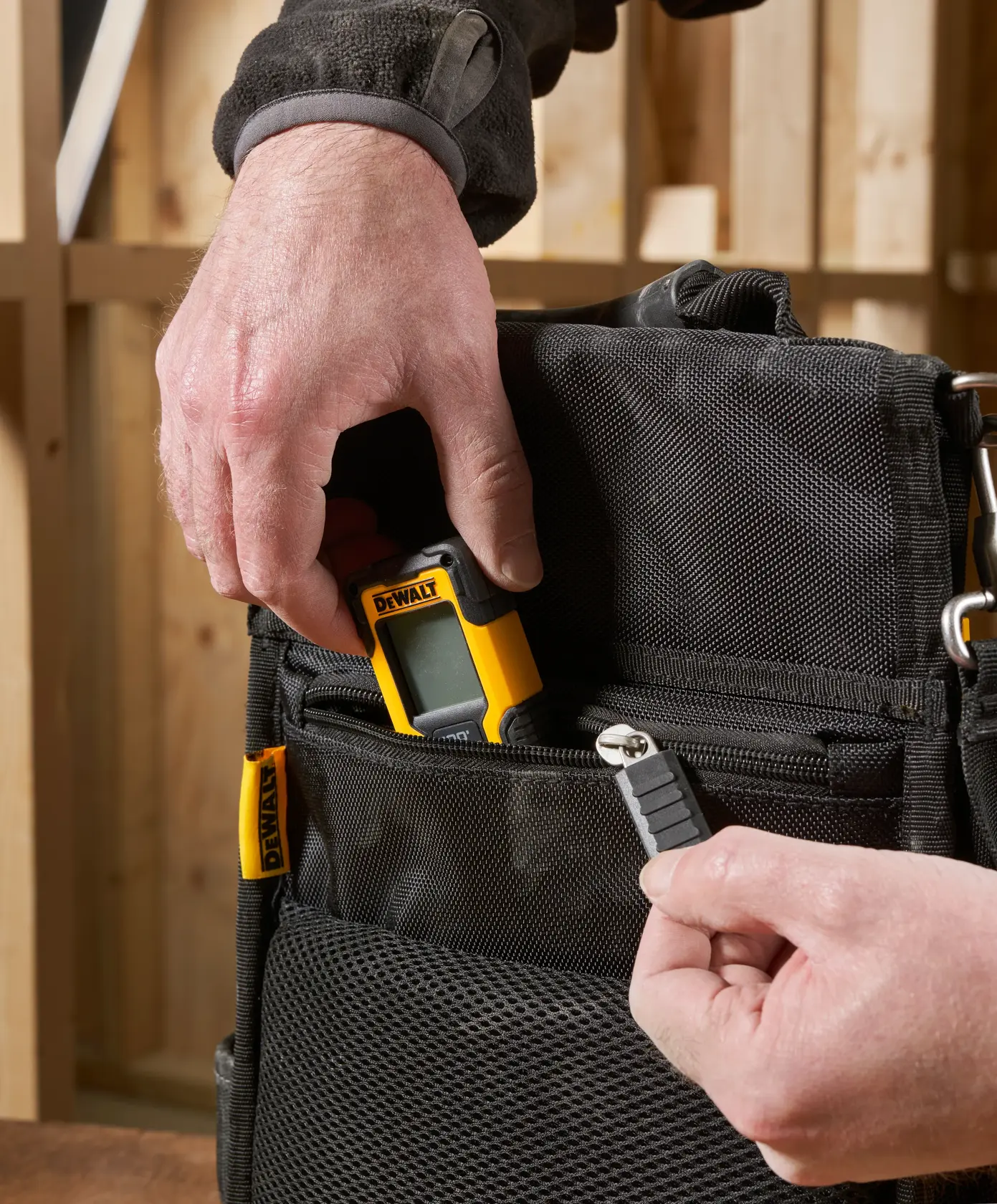 Mão coloca aparelho DEWALT e bit em bolsa preta com zíper em ambiente de oficina.
