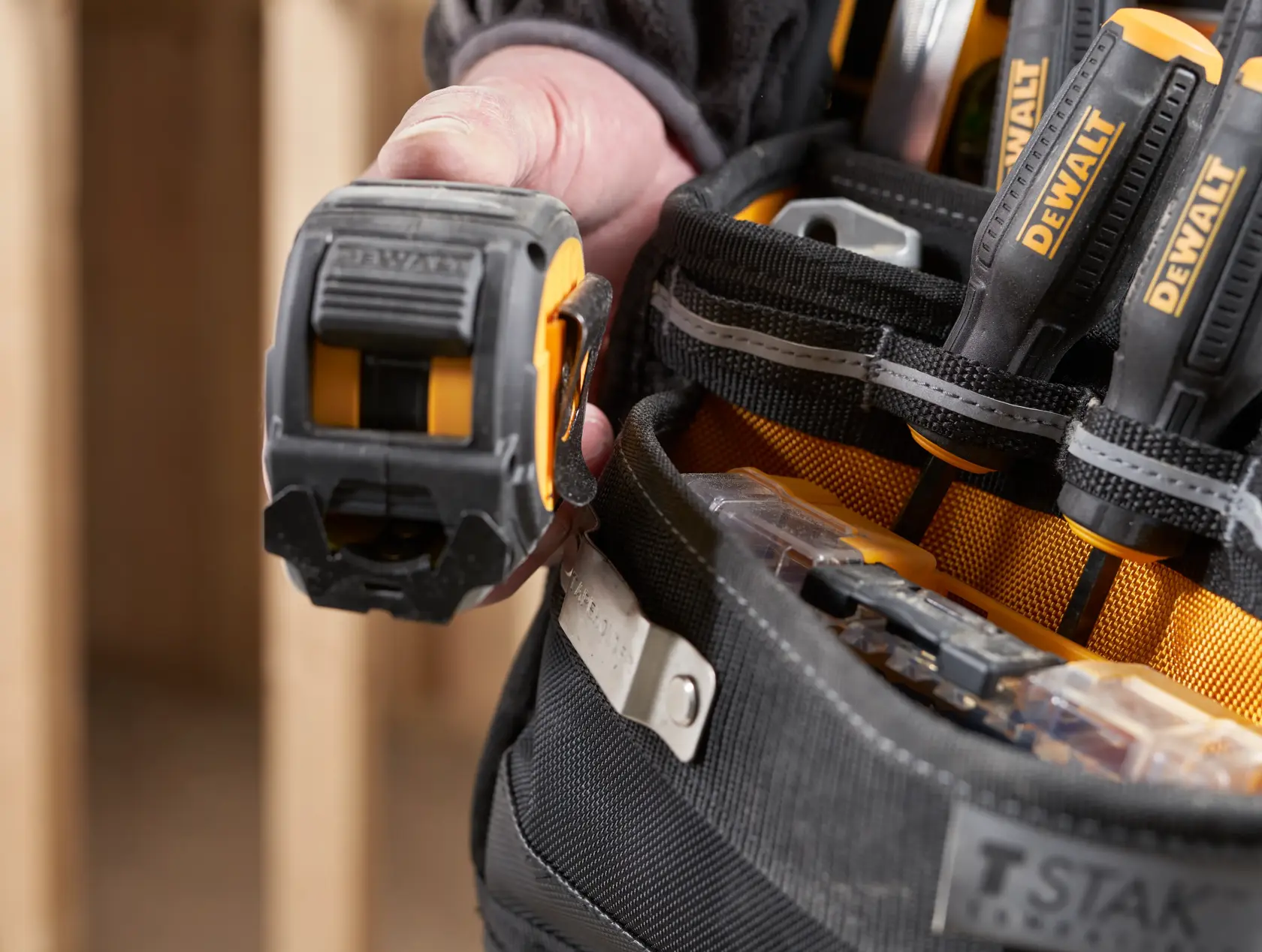 Close-up de um cinto DEWALT com mão segurando fita métrica e chaves de fenda nos bolsos.
